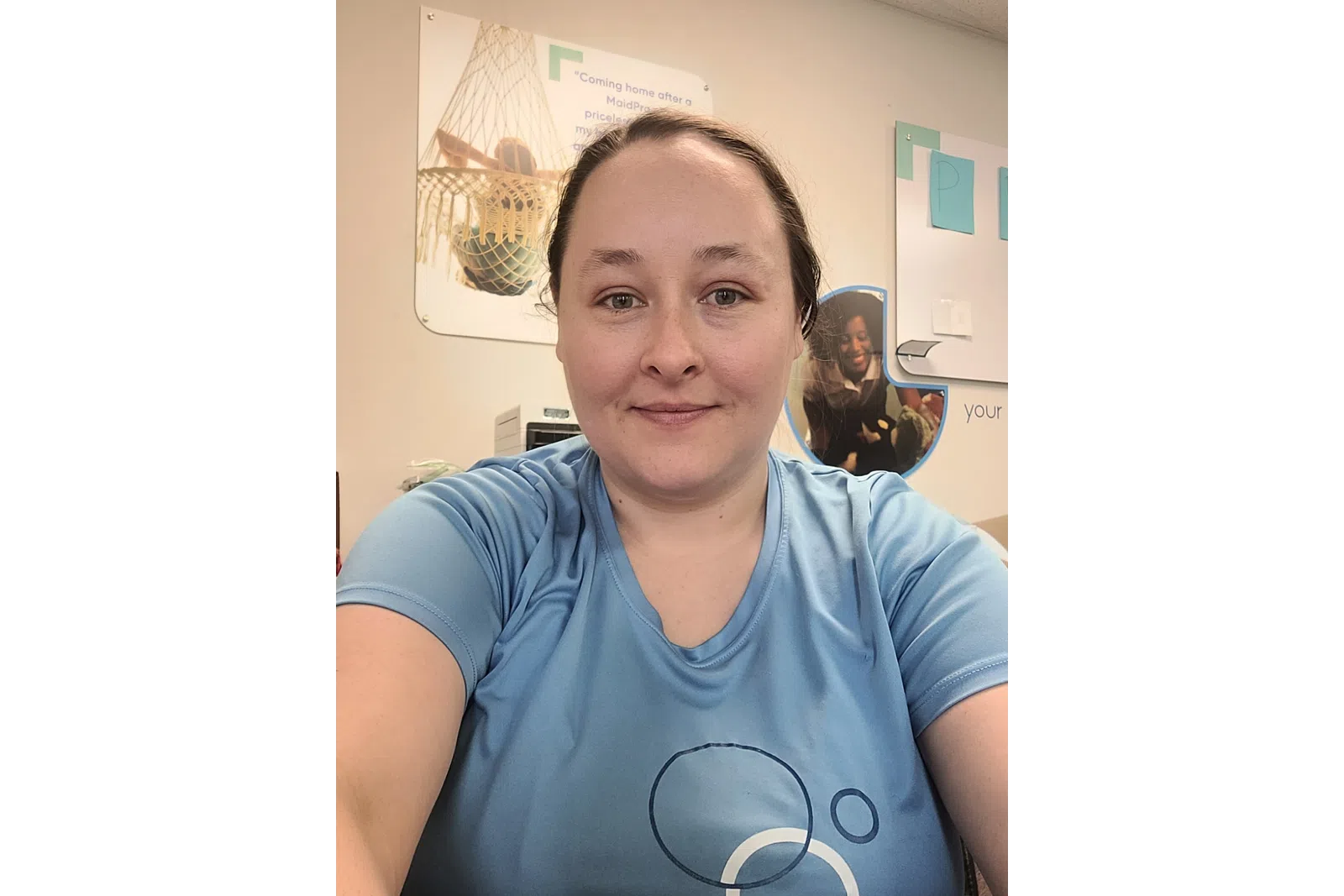 Smiling woman in light blue shirt takes a selfie in an office with posters and papers on the wall behind her.