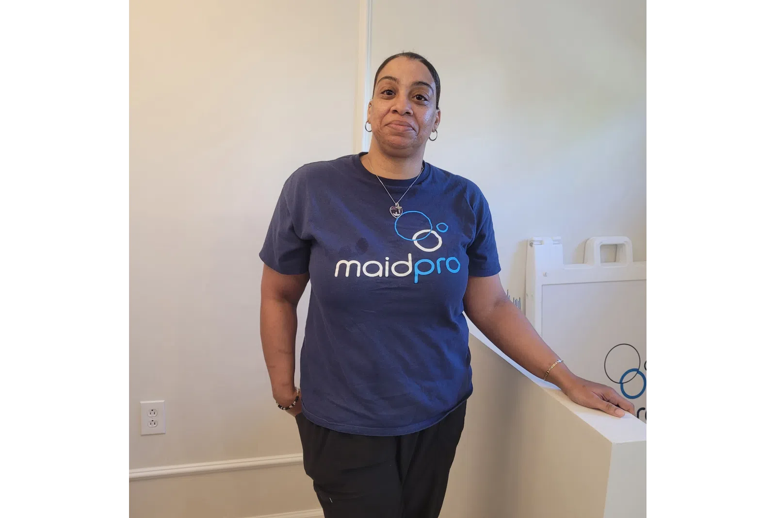 Woman in a navy MaidPro shirt standing indoors beside a white partition with cleaning supplies visible.