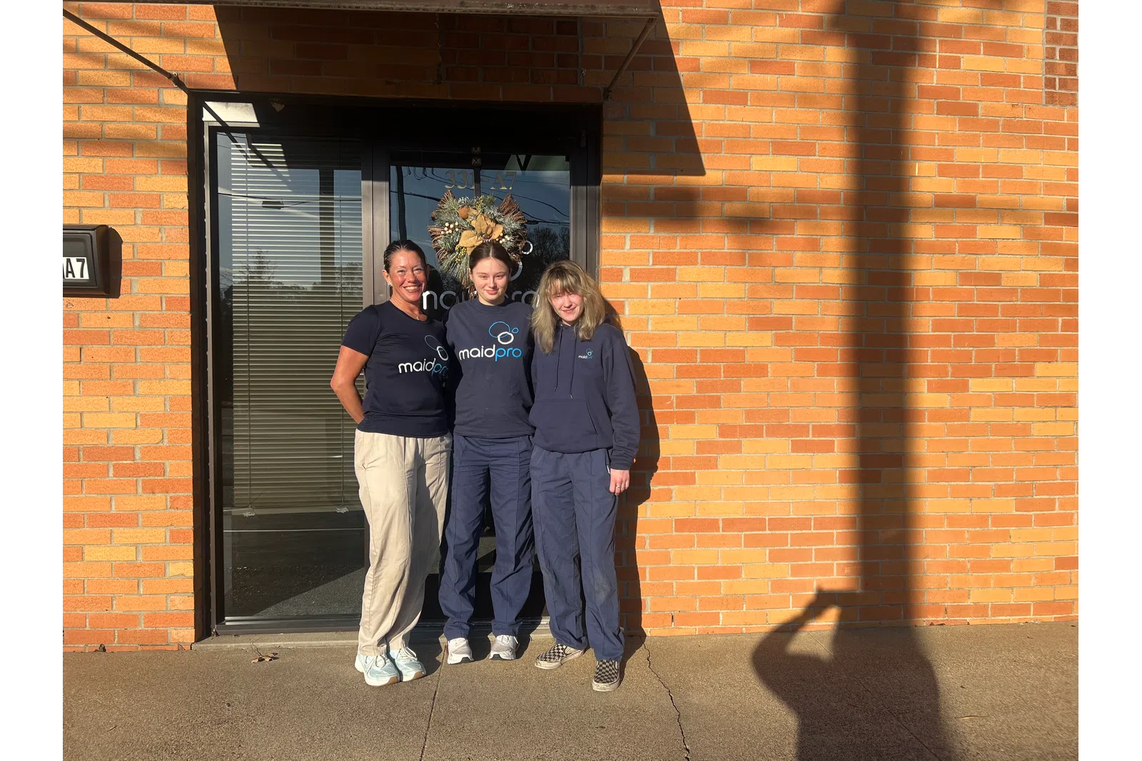 Three women in MaidPro uniforms standing outside a brick building entrance with a wreath on the door.