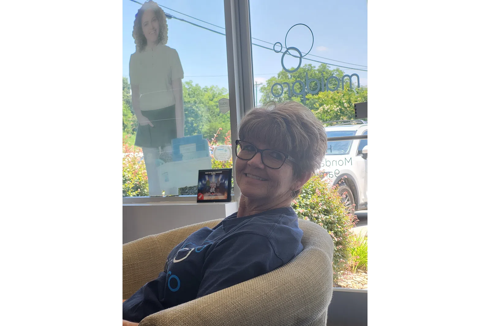 Smiling woman wearing glasses sitting in a chair near a window with outdoor greenery and a standing cutout figure visible.
