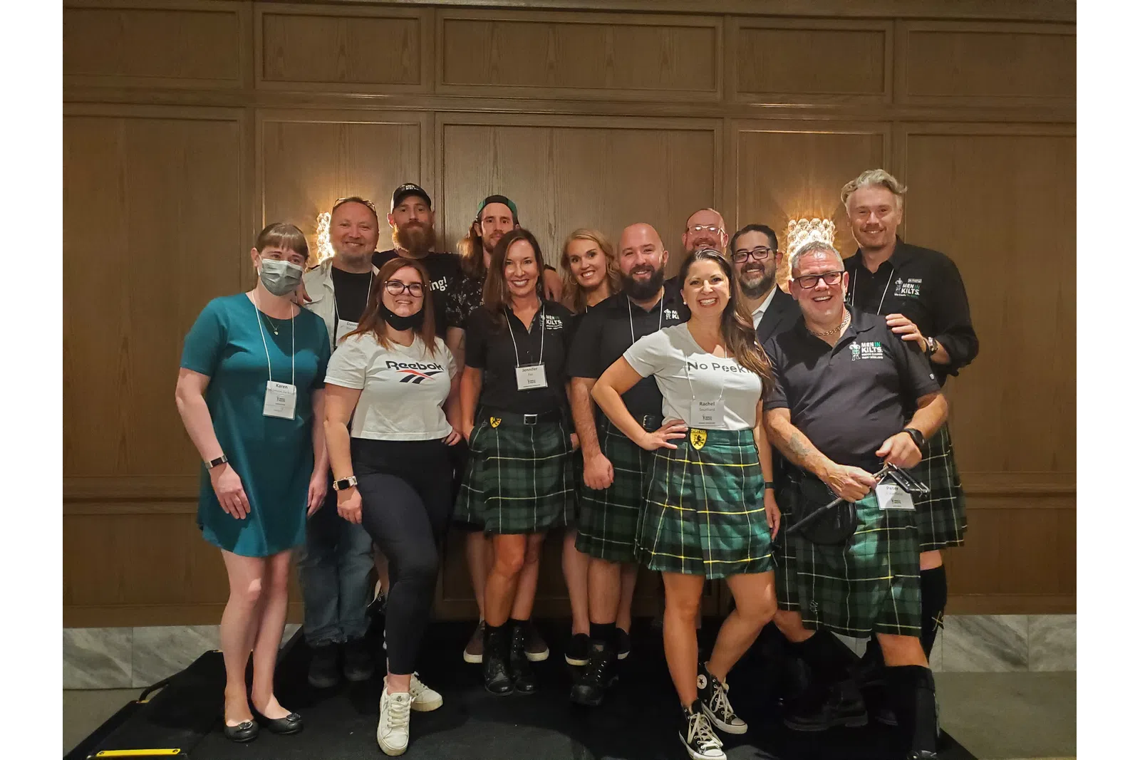 Group of eleven people smiling indoors, most wearing green tartan kilts and casual tops, posing in front of wood panel wall.