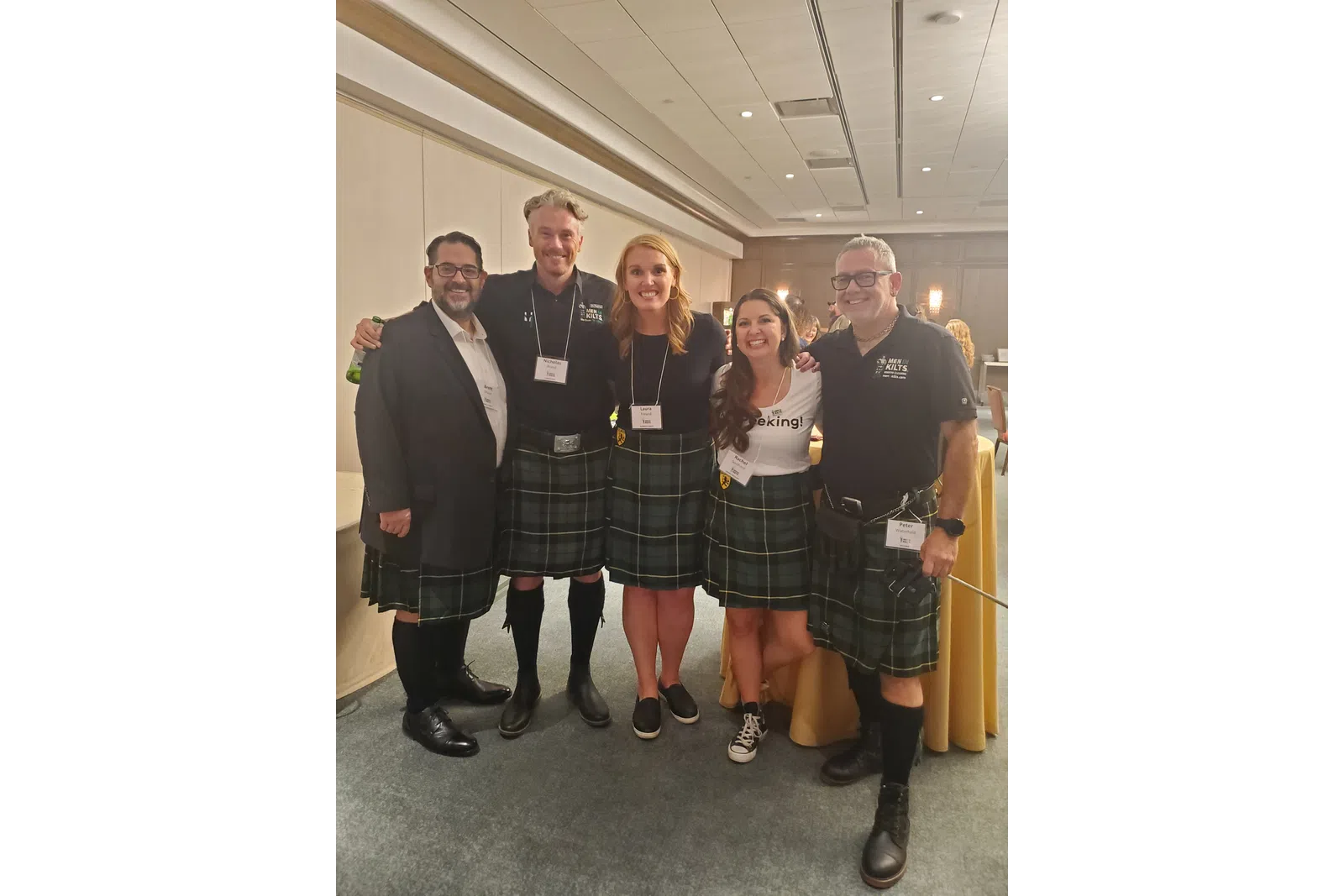 Five people smiling indoors wearing matching green tartan kilts and name badges at an event.