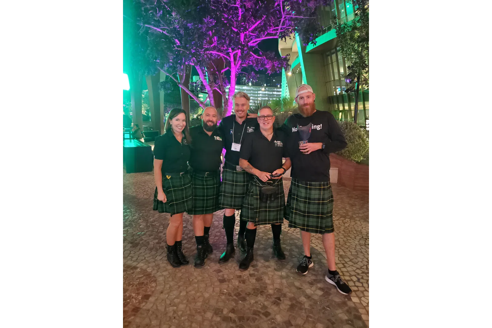 Group of five people wearing green plaid kilts and black tops posing outdoors at night with colorful tree lighting.