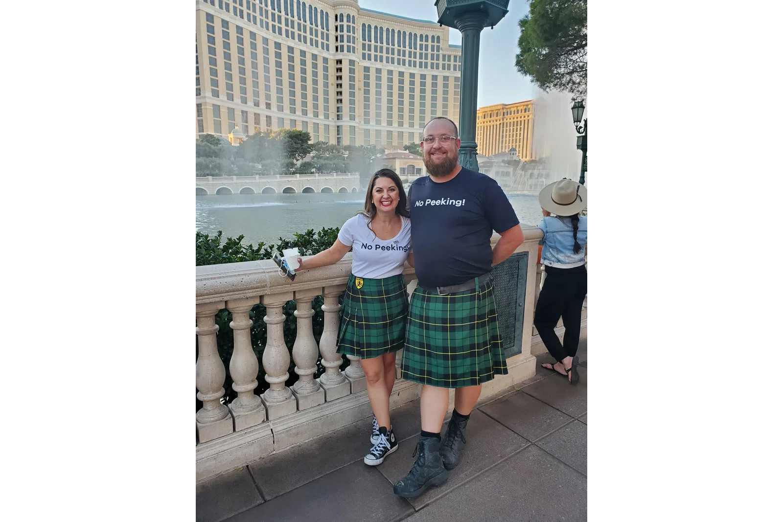 Smiling couple wearing matching green plaid kilts and No Peeking shirts by a fountain with hotel buildings in the background