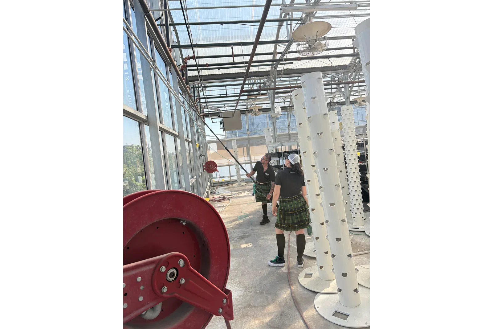 Two people in kilts inside a greenhouse with vertical hydroponic towers and red hose reel.
