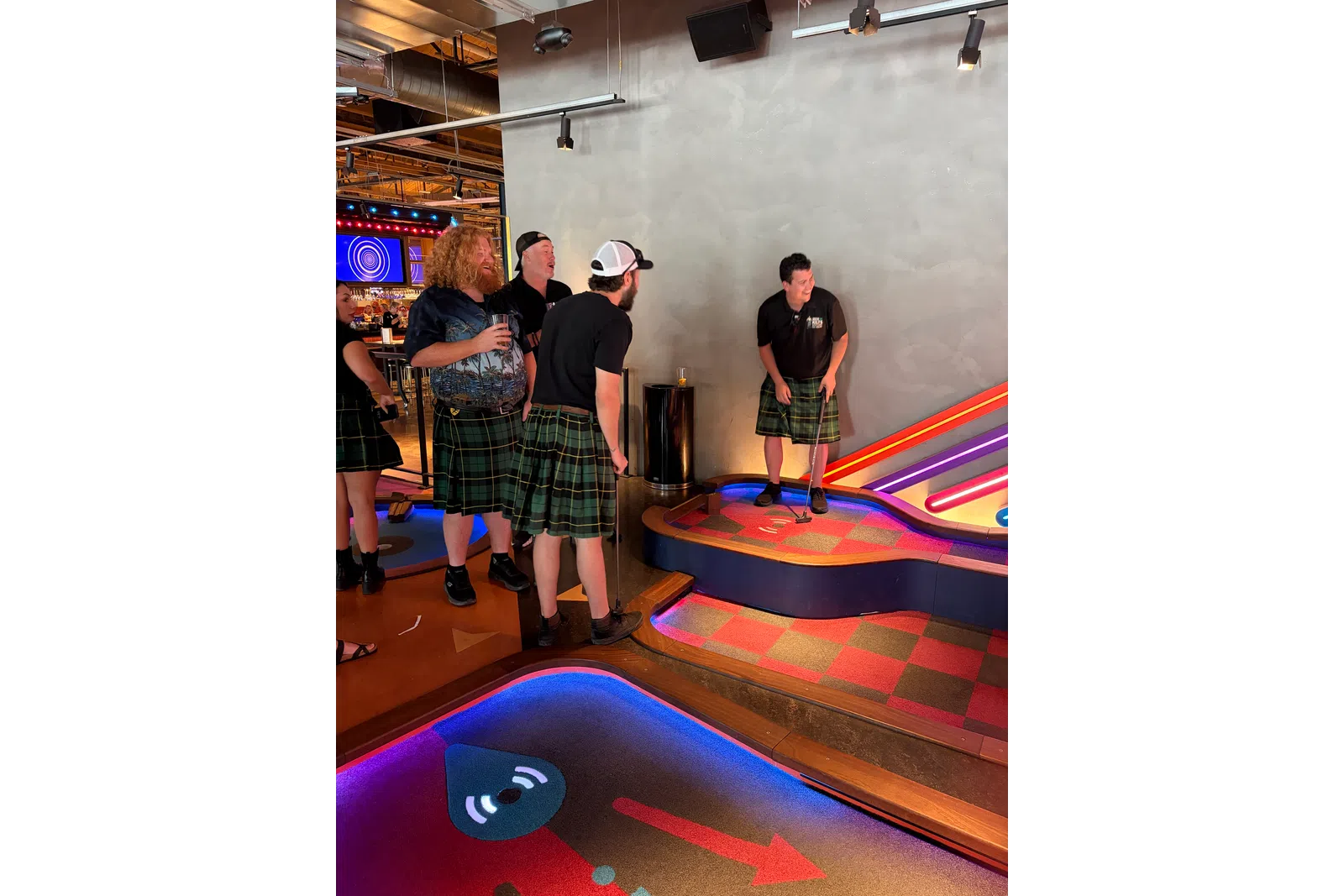 Group of adults wearing kilts playing indoor mini golf in a colorful, modern entertainment venue.