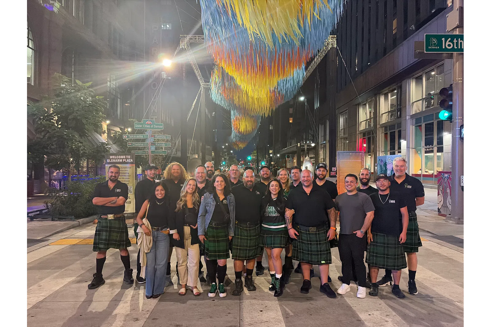 Group of people wearing kilts standing on a city street at night under colorful hanging art installation.