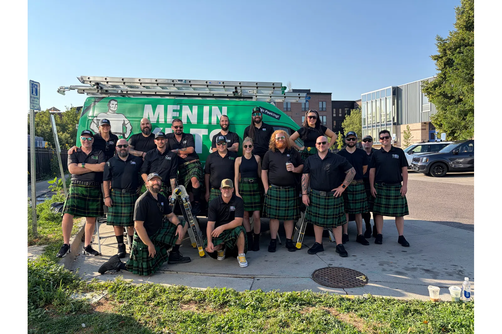 Group of men and women wearing matching black shirts and green kilts posing outdoors by a green work van.