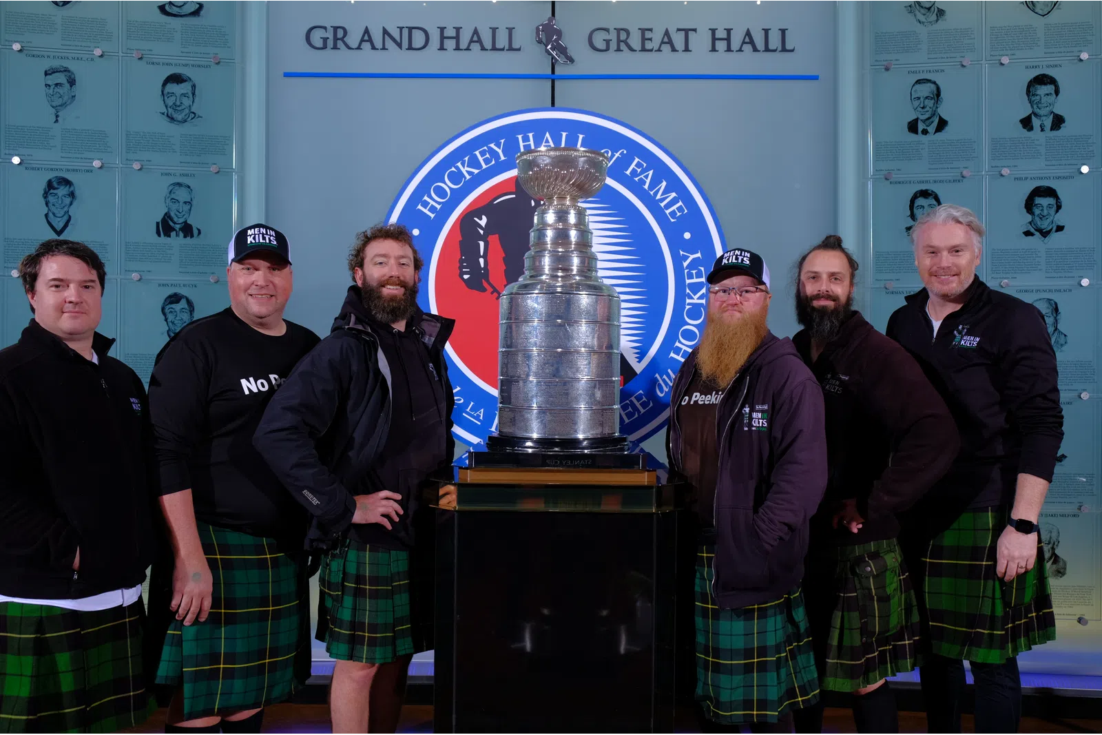 Group of men in green plaid kilts posing around the Stanley Cup in the Hockey Hall of Fame Grand Hall.