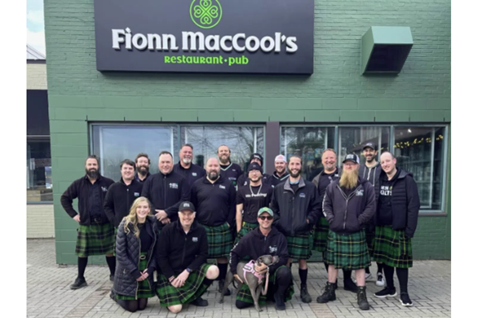 Group of people wearing green plaid kilts posing outside Fionn MacCool's restaurant and pub on a city sidewalk.