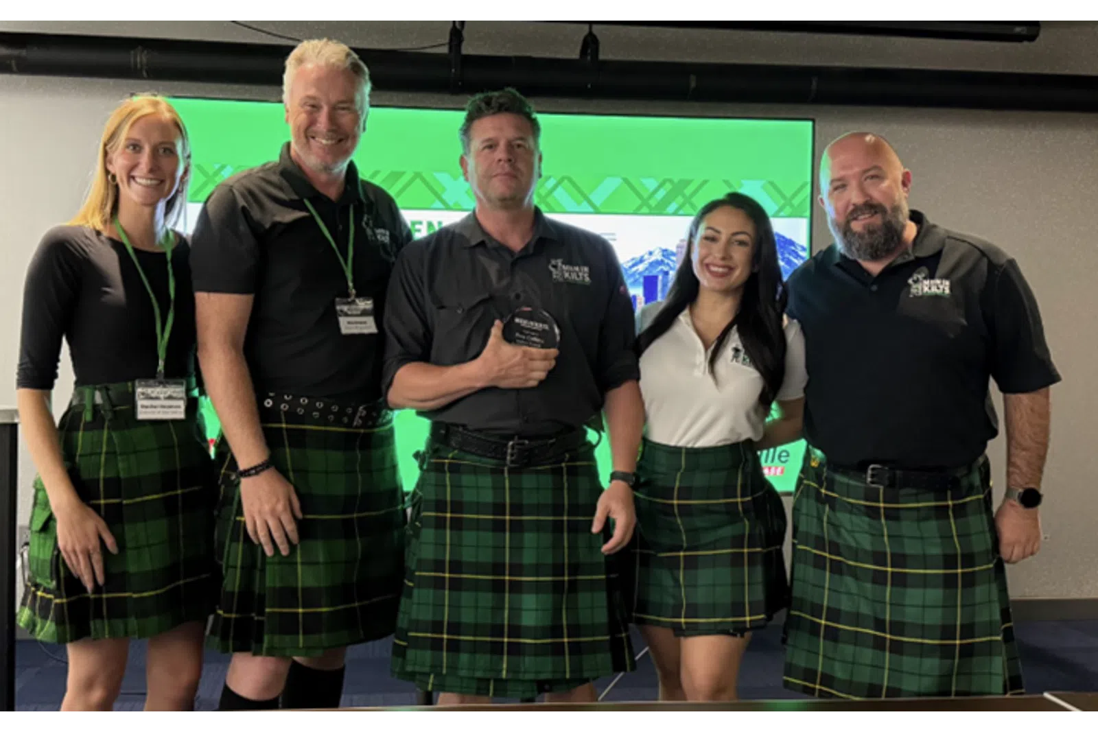 Five people wearing green plaid kilts posing together indoors at an event with a green screen behind them.