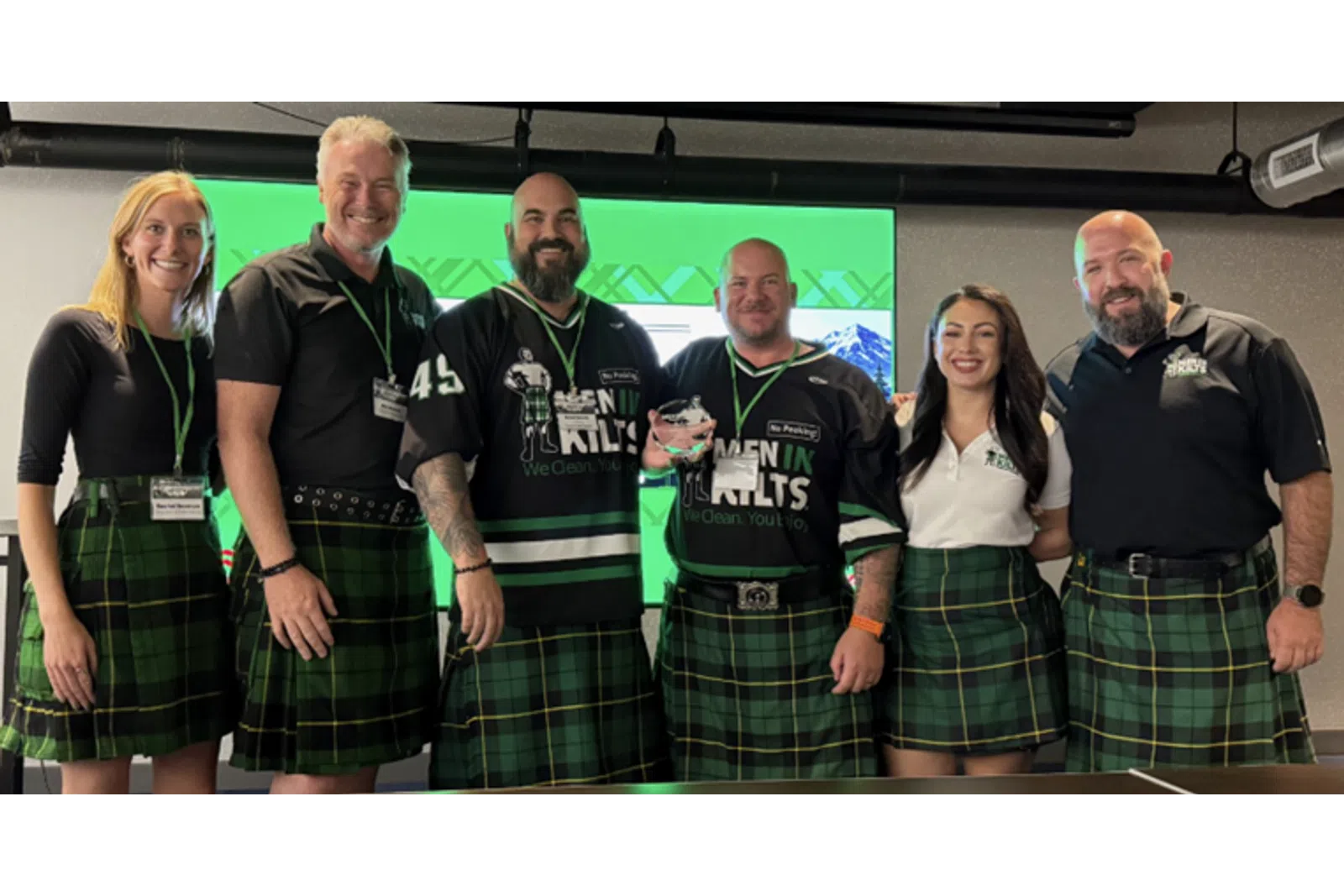 Group of six people wearing green plaid kilts and black shirts, smiling in front of a green screen backdrop.