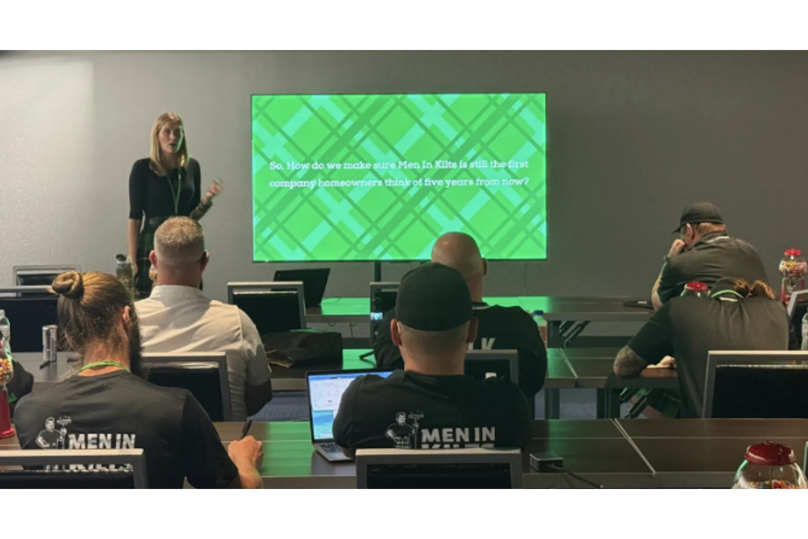 Presenter giving a presentation to seated attendees with a green slide about Men In Kilts company future vision.