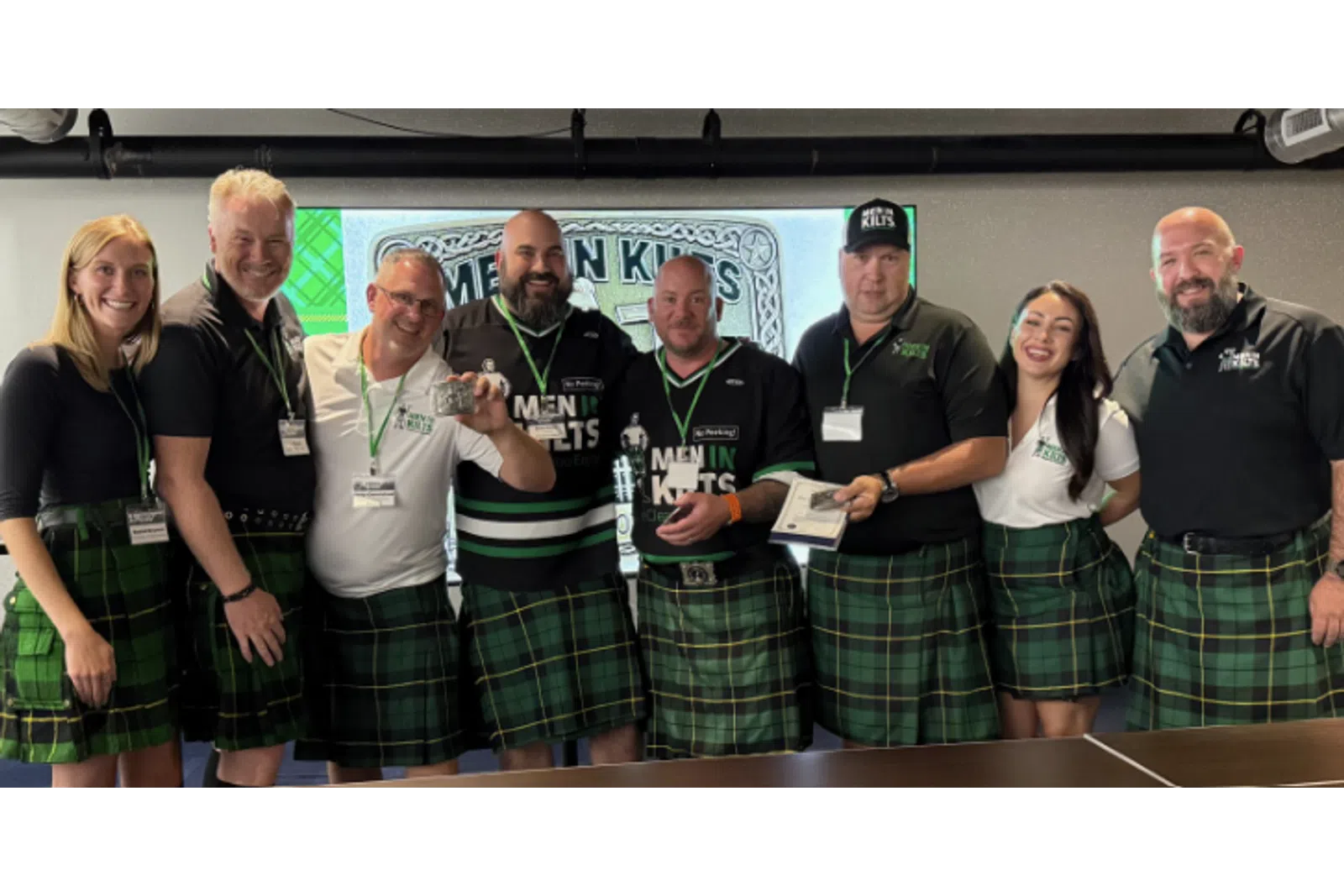 Group of men and women wearing green tartan kilts posing at a Men in Kilts event with smiles and awards.