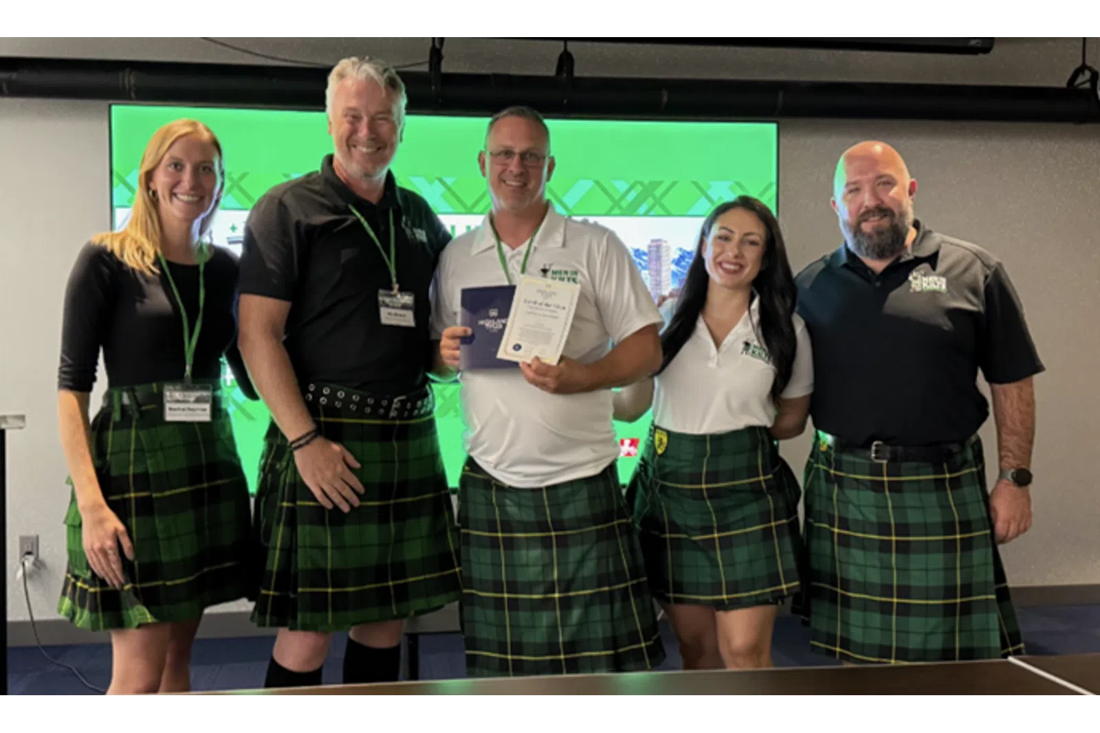 Group of five people wearing green plaid kilts, smiling and holding certificates in front of a green screen.