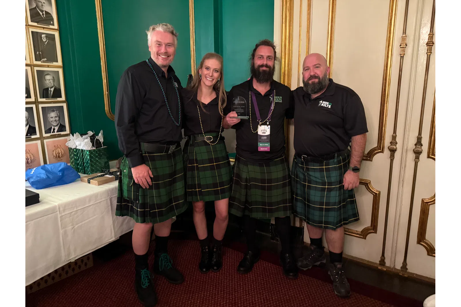 Four people wearing green plaid kilts posing indoors, smiling and holding an award, standing in ornate room.
