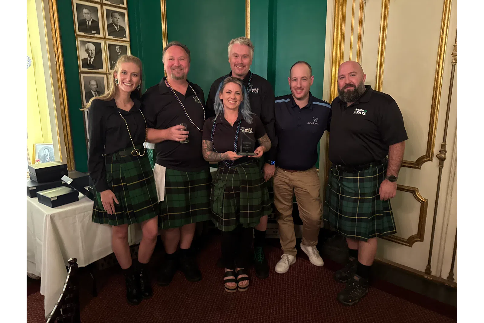 Group of six people, five wearing green tartan kilts, posing indoors with award in decorated room.