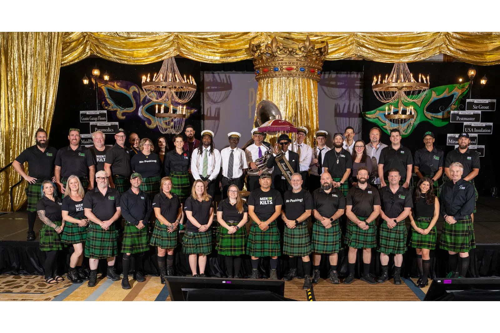 Group photo of men and women wearing green plaid kilts and black shirts posing on stage under gold curtains and chandeliers.
