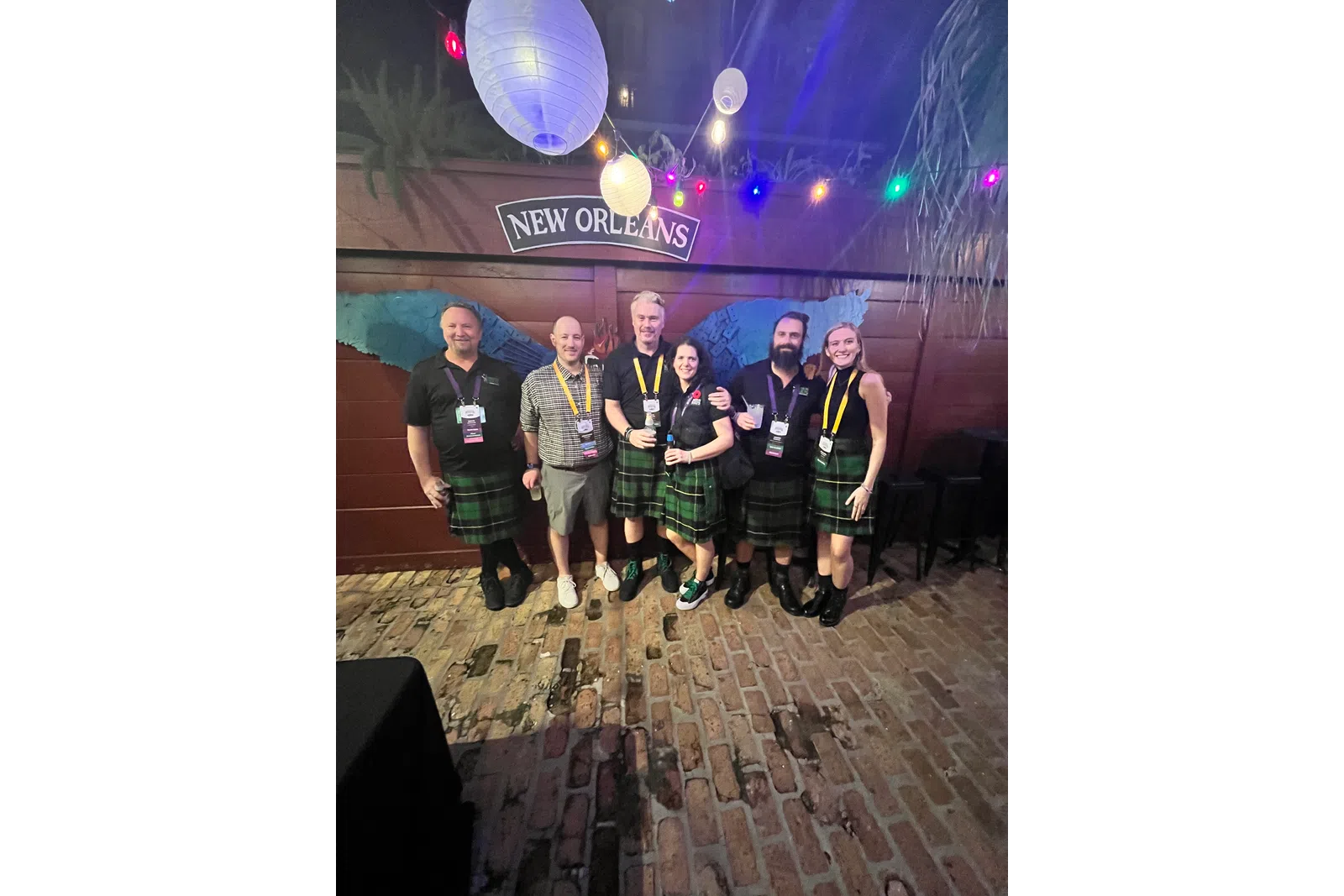 Group of six people wearing kilts at a New Orleans venue with hanging lanterns and colorful string lights