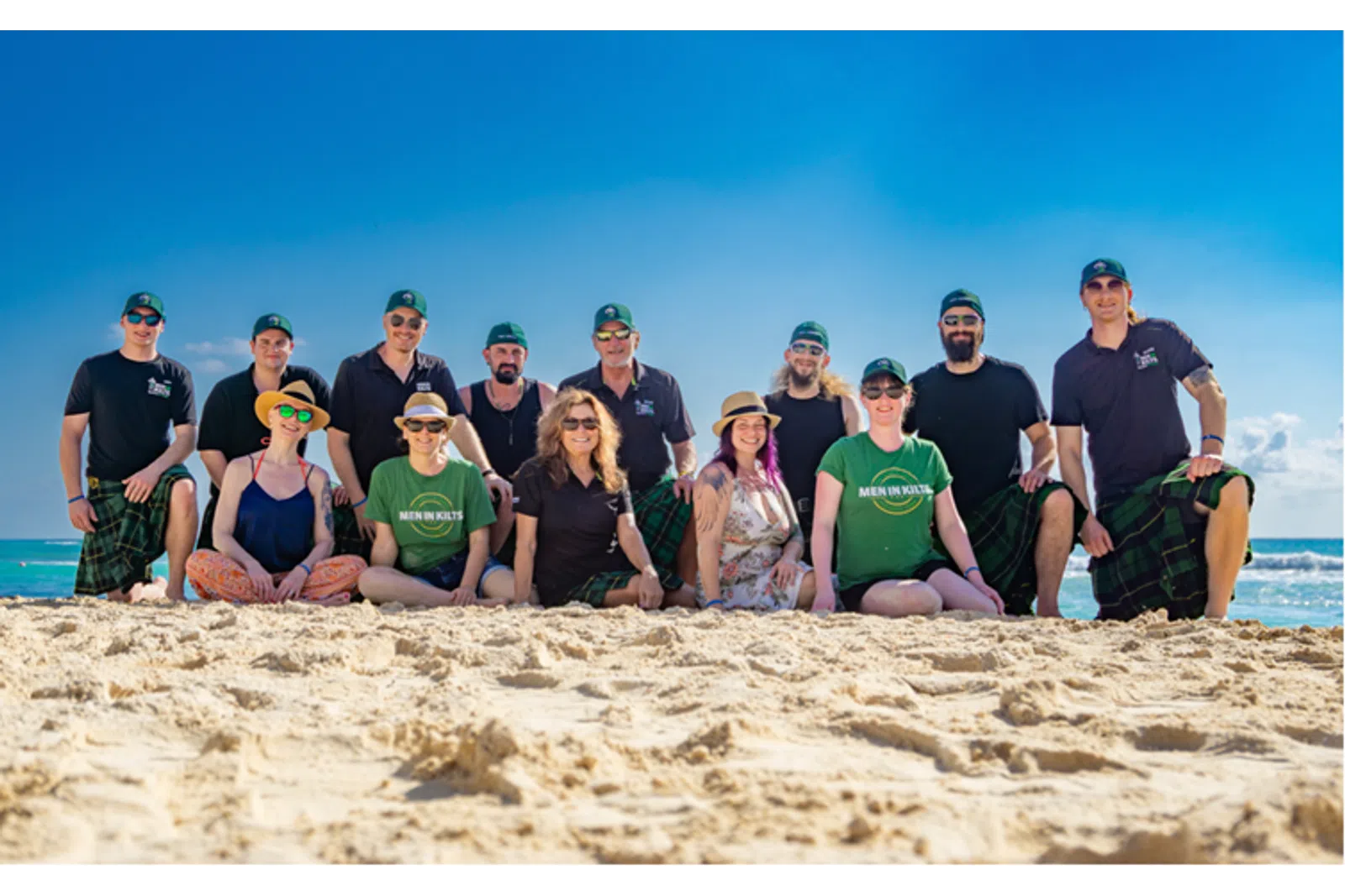 Group of people in kilts and casual wear posing on sunny beach with blue sky and ocean in background