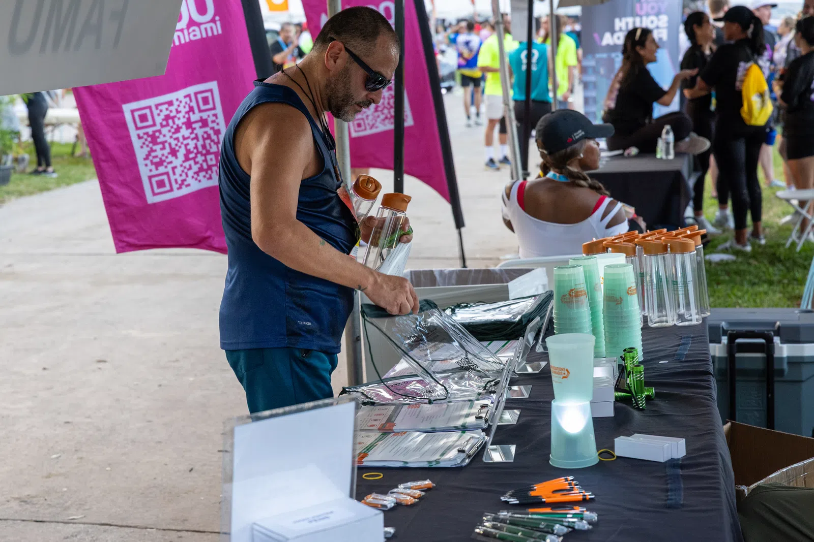 Man in sunglasses browsing items on a table with water bottles, cups, pens at an outdoor event booth.