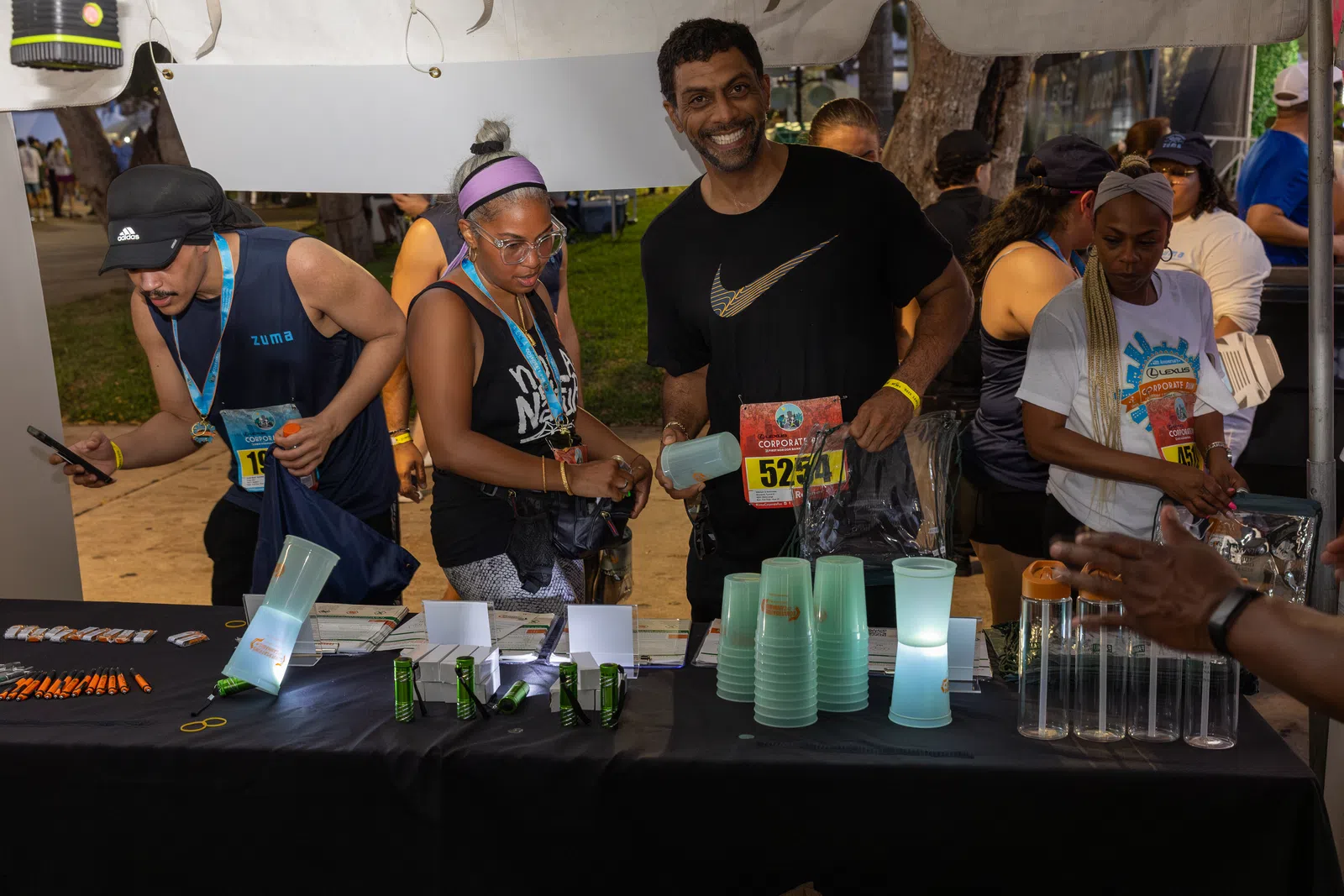 Participants at a race event booth collecting water cups and race bibs under a tent at dusk.