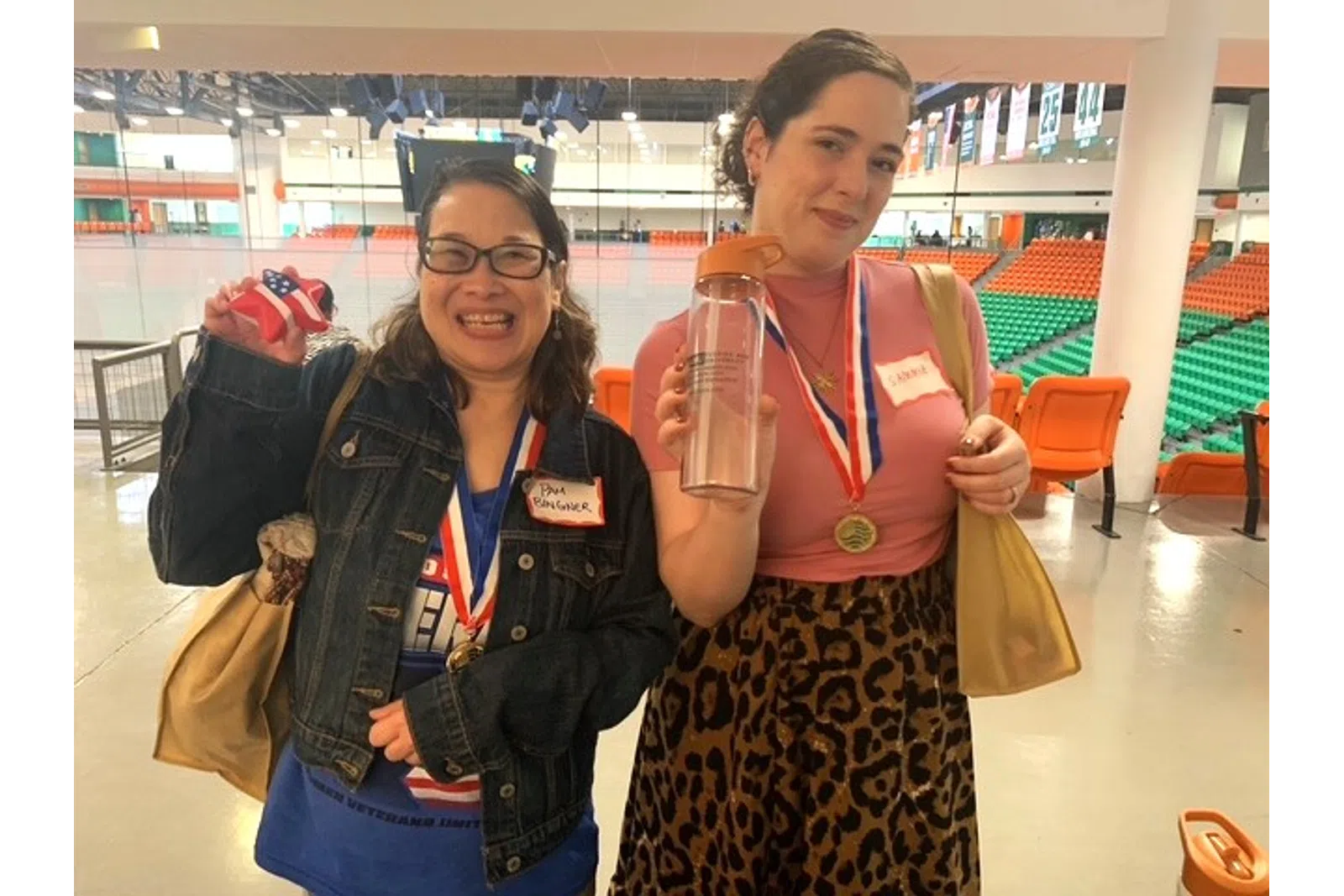 Two women wearing medals smile and hold trophies inside an indoor sports arena with orange and green seats.