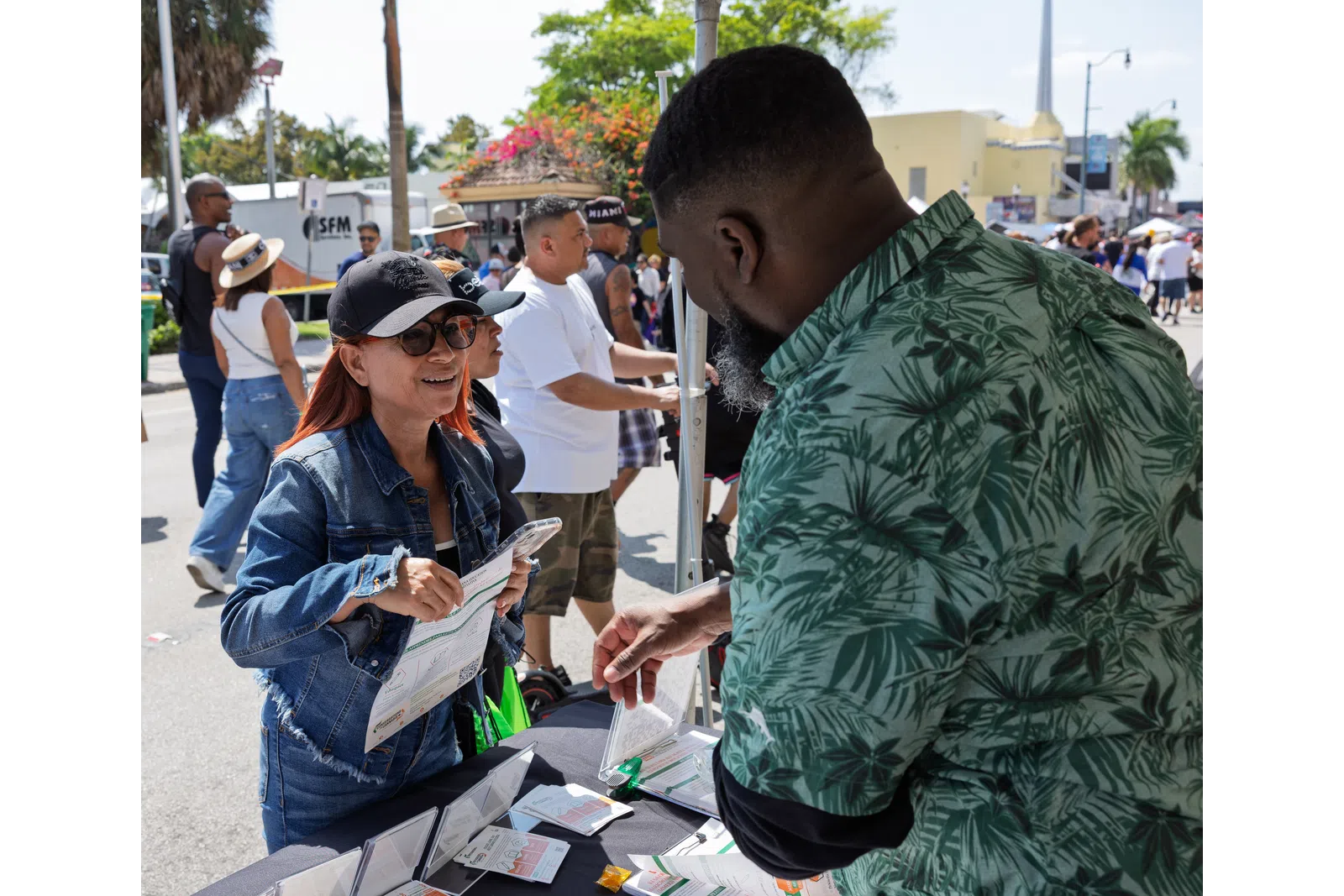 Woman in denim jacket and cap engaging with man in green shirt at outdoor event booth on sunny day