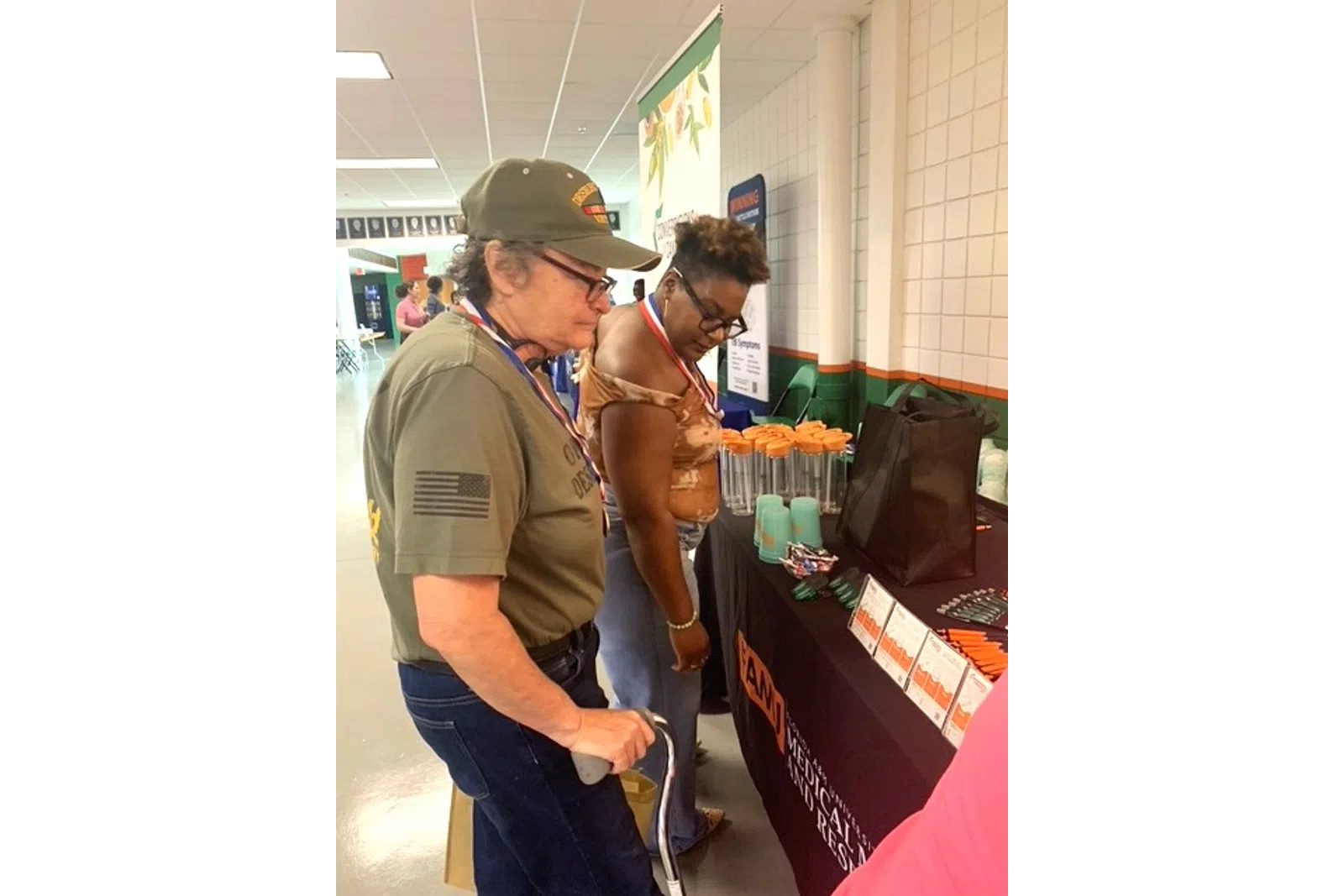 Two women examining promotional items on a table at an indoor community event or expo.