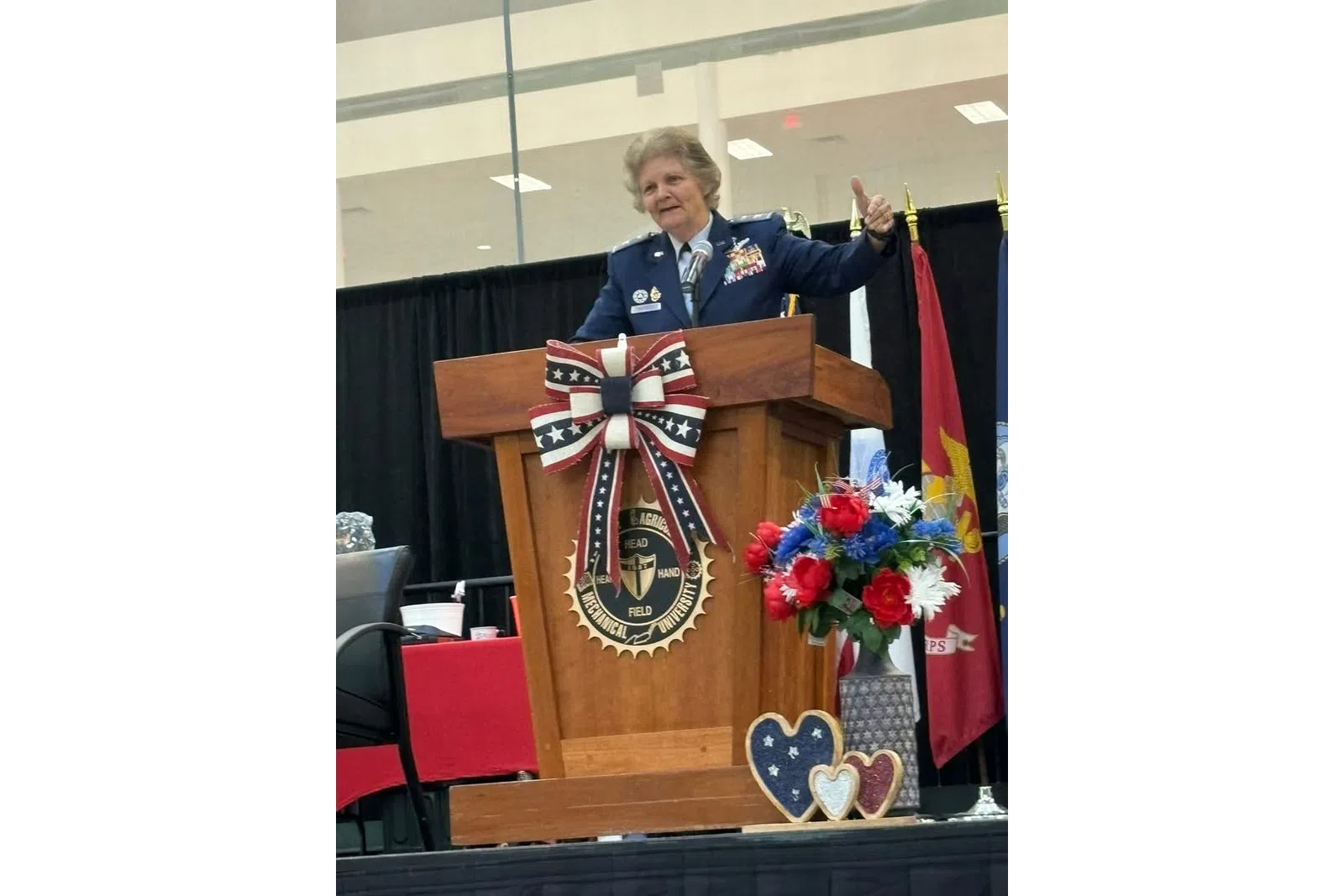 Female military officer in uniform giving speech at decorated wooden podium with patriotic decor and flags.
