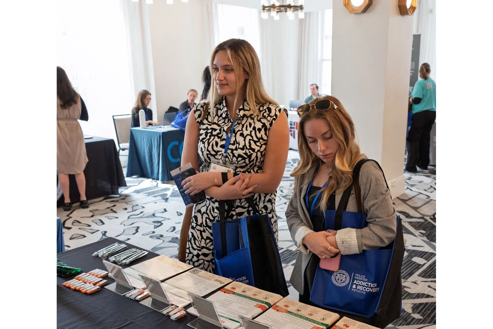 Two women with blue tote bags browsing informational brochures at an indoor event or conference table.