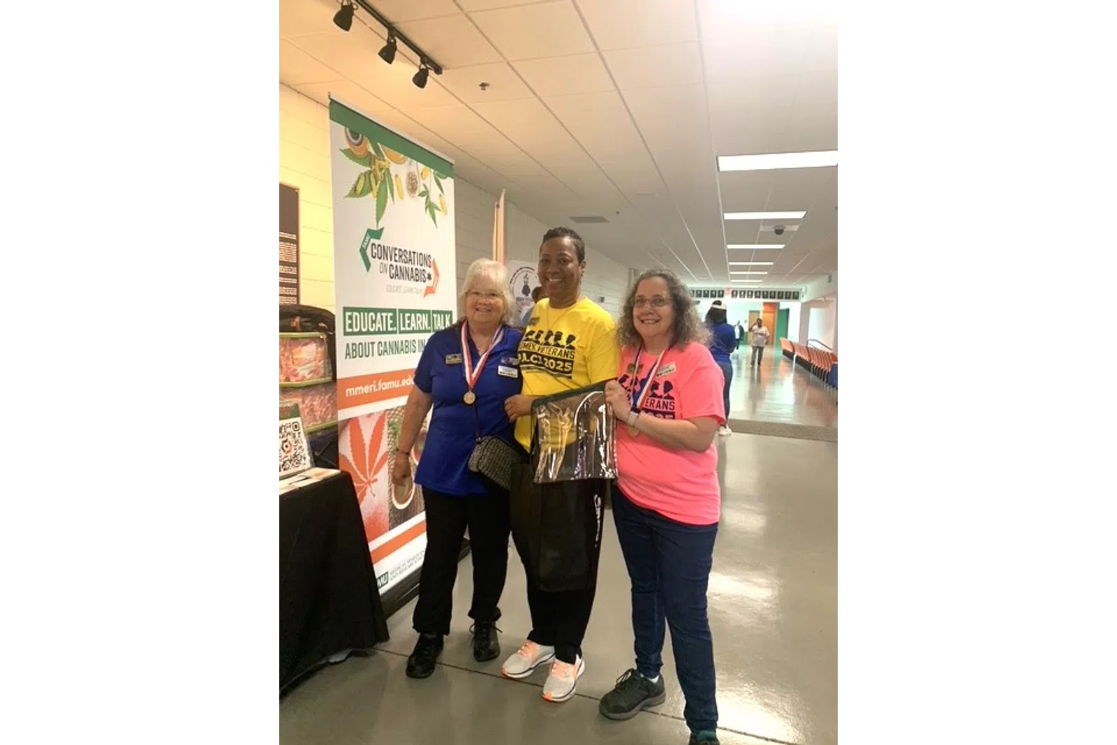 Three smiling women stand indoors at a cannabis education event with medals and colorful shirts.
