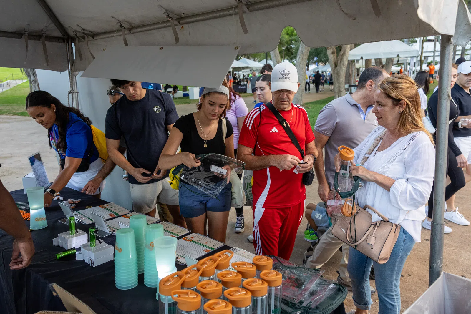 People selecting reusable water bottles under a tent at an outdoor event with trees and tents in the background