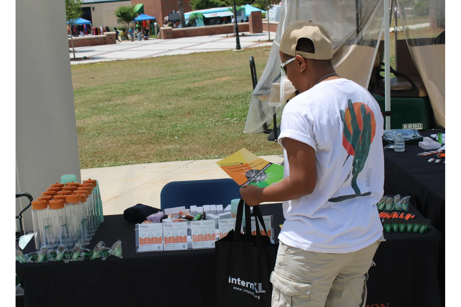 Person browsing an outdoor vendor table with promotional items and brochures on a sunny day.