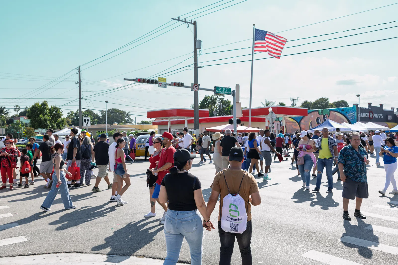 Crowded street festival with diverse people walking, an American flag, and a red canopy in daylight.