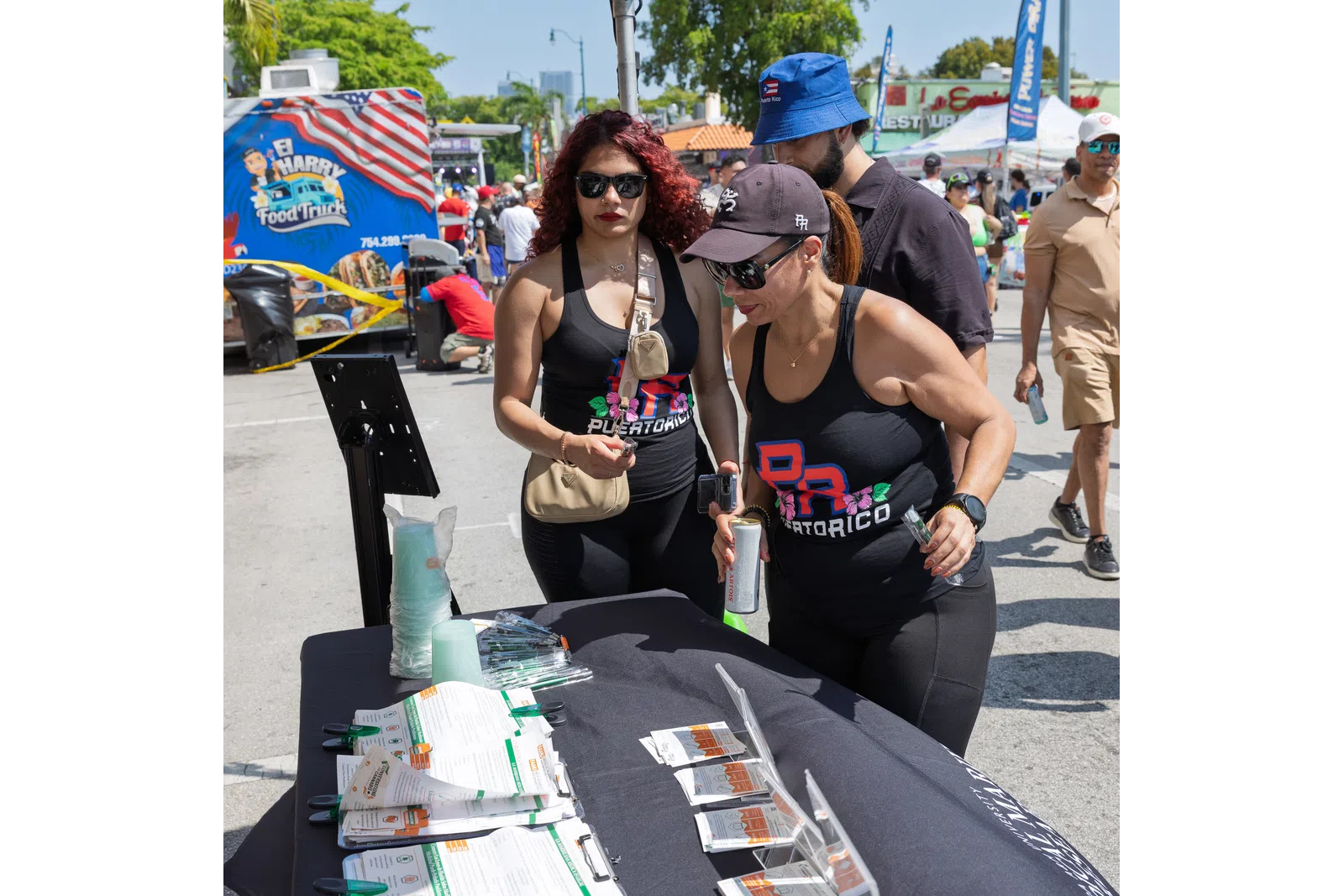 Two women in Puerto Rico tank tops explore products on a table at an outdoor street festival with food trucks.