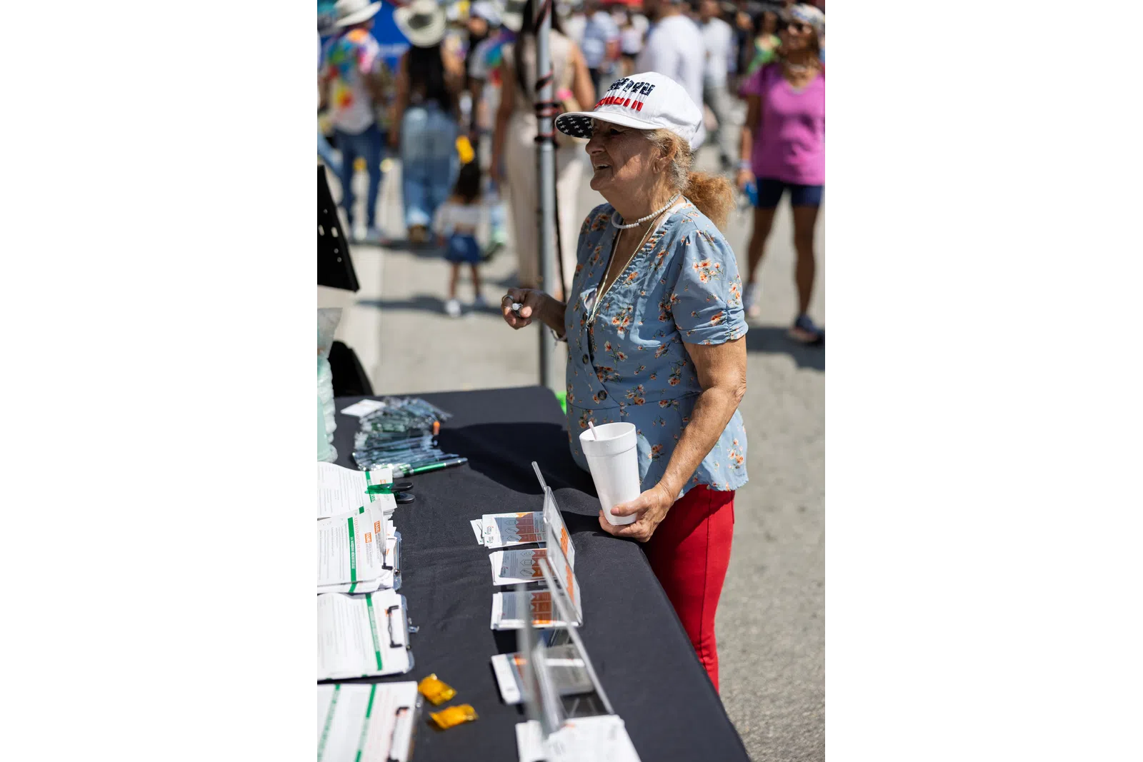 Older woman in a floral shirt and white hat stands at an outdoor information table holding a white cup.