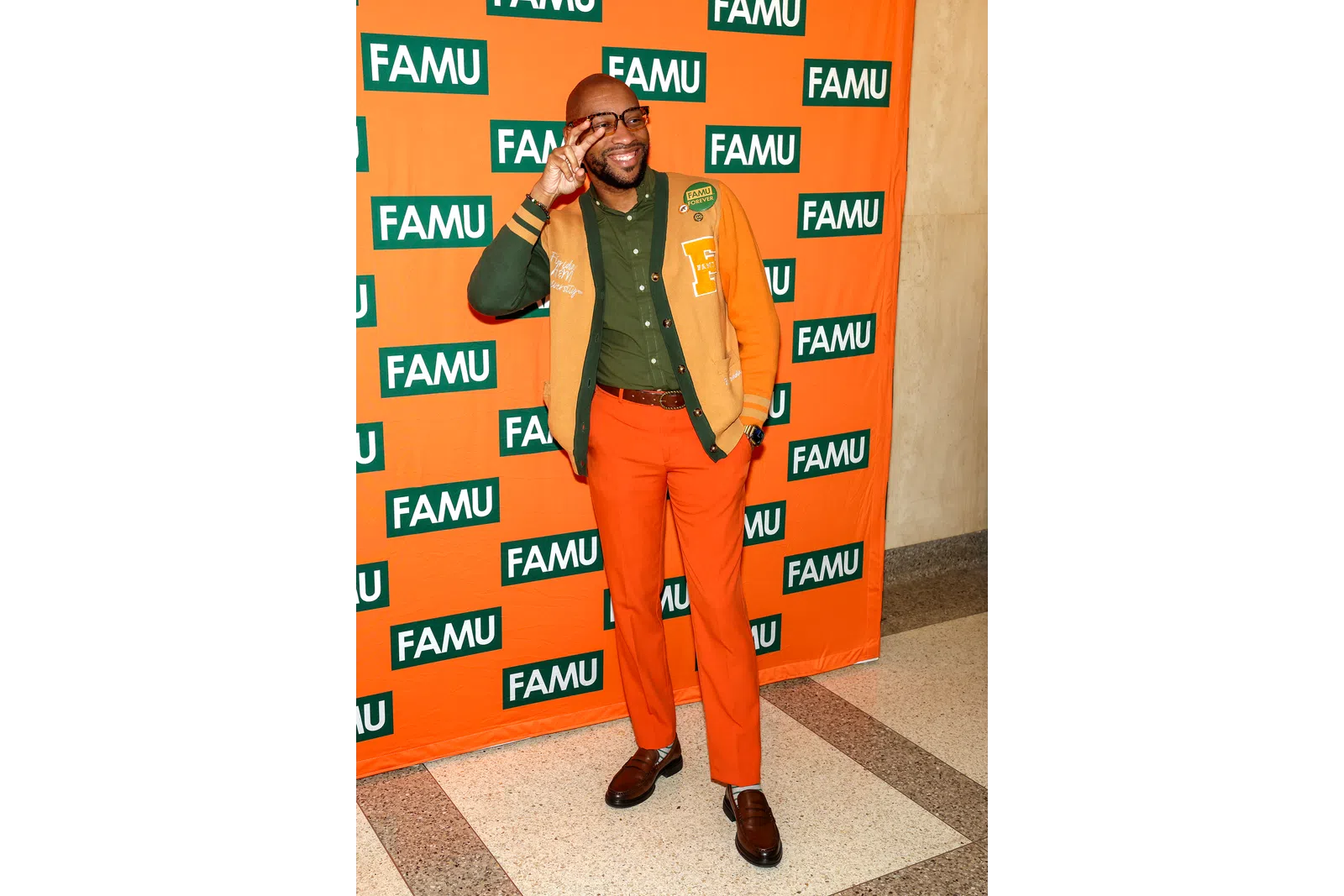 Man in orange and green outfit posing joyfully with FAMU backdrop at an indoor event