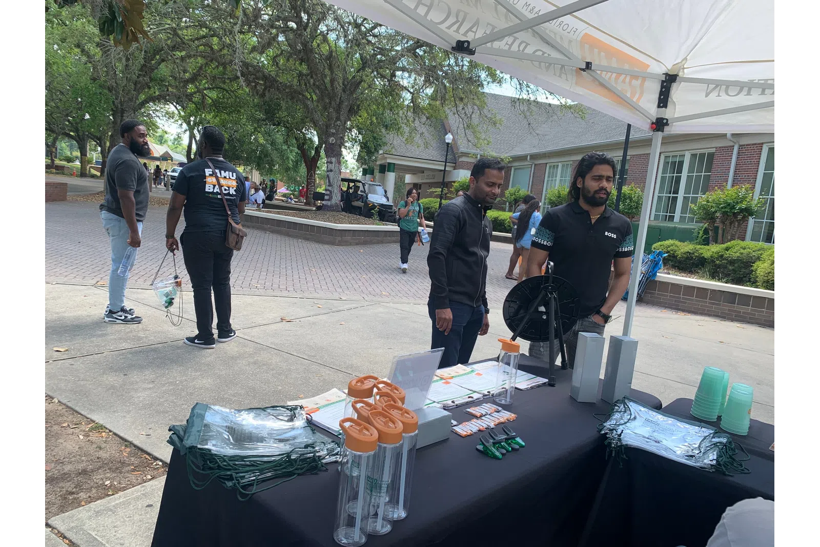 Outdoor campus event with information booth offering promotional items and two men staffing the table under a canopy.