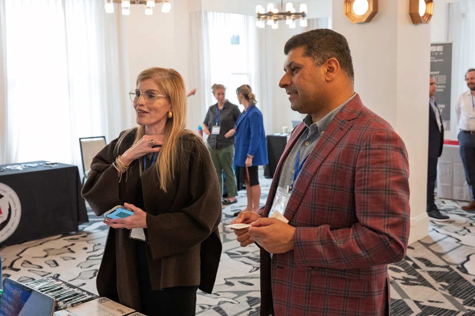 Two professionals talking at a business networking event in a well-lit conference room with several attendees in the
