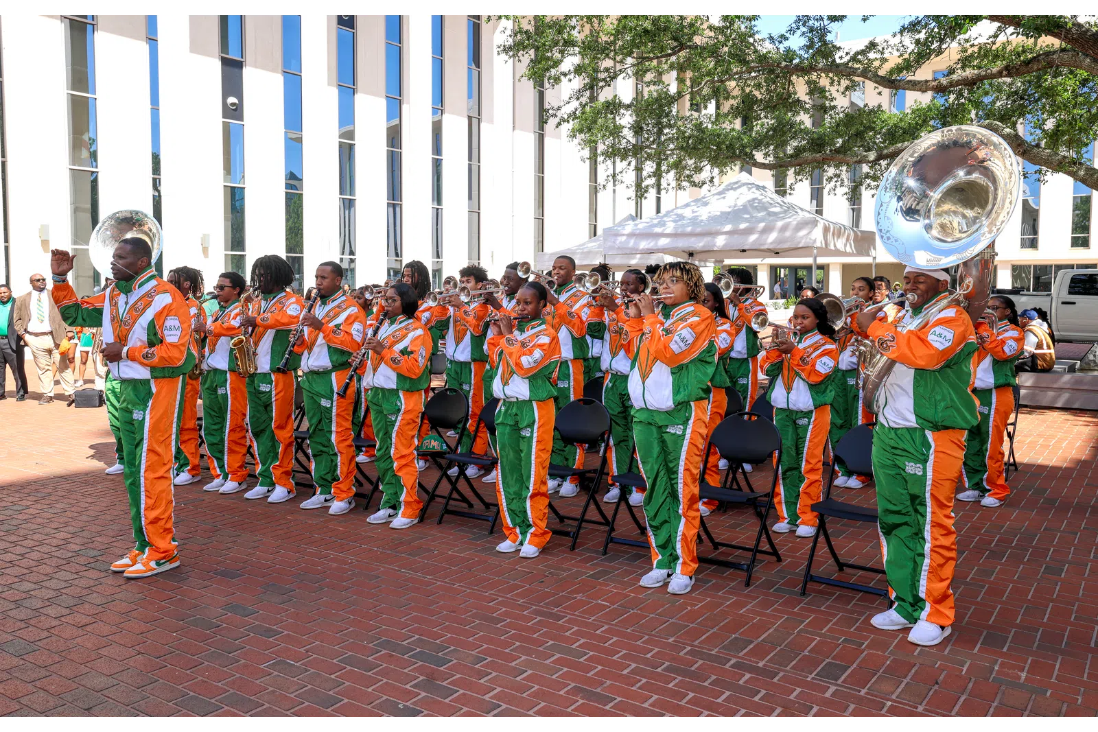 Marching band in orange and green uniforms playing instruments outdoors in front of a building under sunlight.