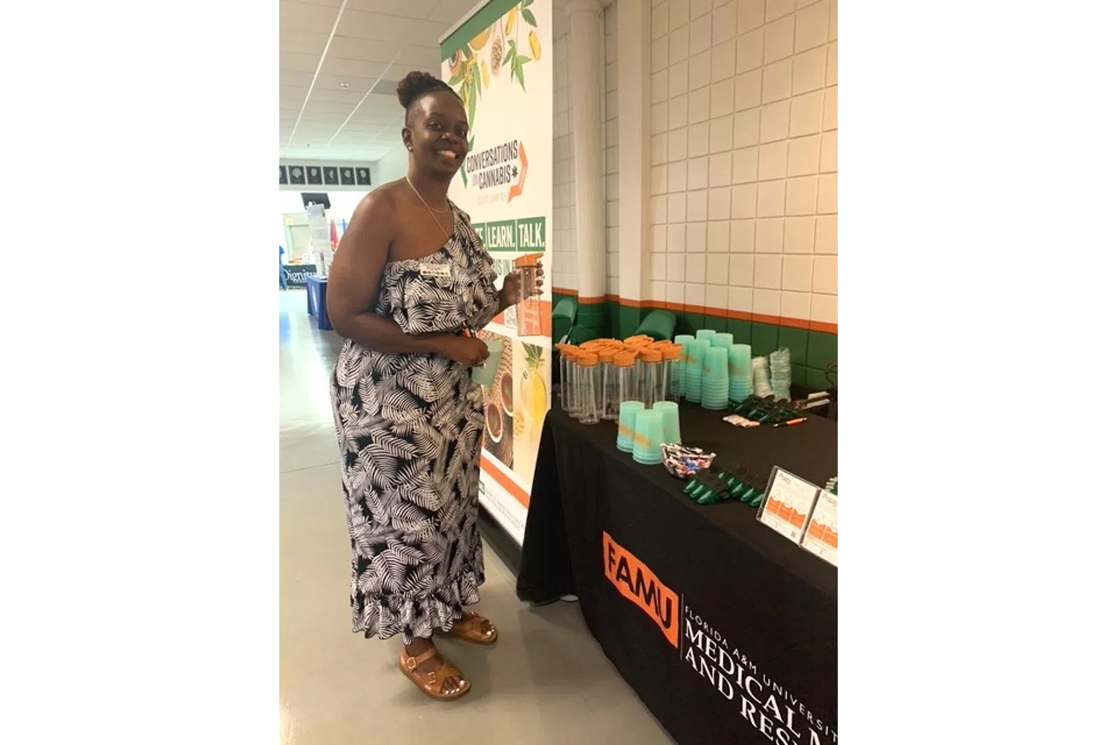 Woman in patterned dress stands by FAMU table with promotional cups and educational cannabis awareness banner.