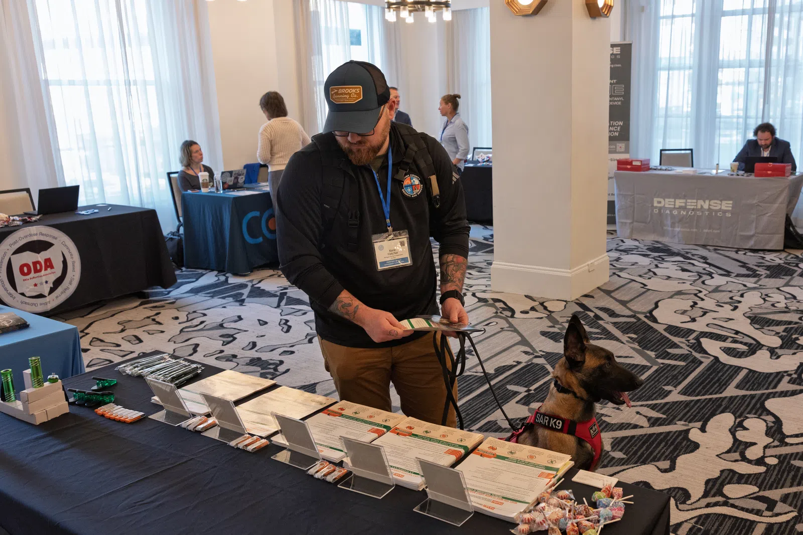 Man with badge and cap stands at exhibitor table reading brochures with a SAR K9 dog beside him inside a conference hall.