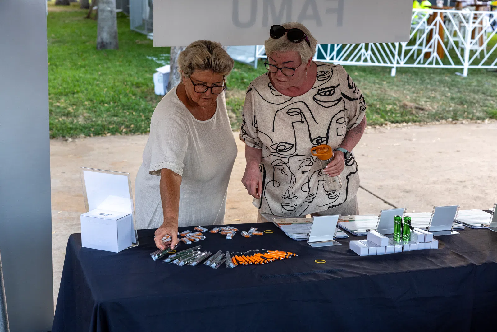 Two elderly women examining promotional items like pens and keychains on a black table at an outdoor event booth.