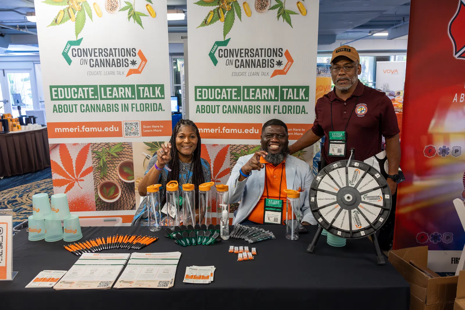 Three people at a cannabis education booth with informational banners, promotional items, and a prize wheel