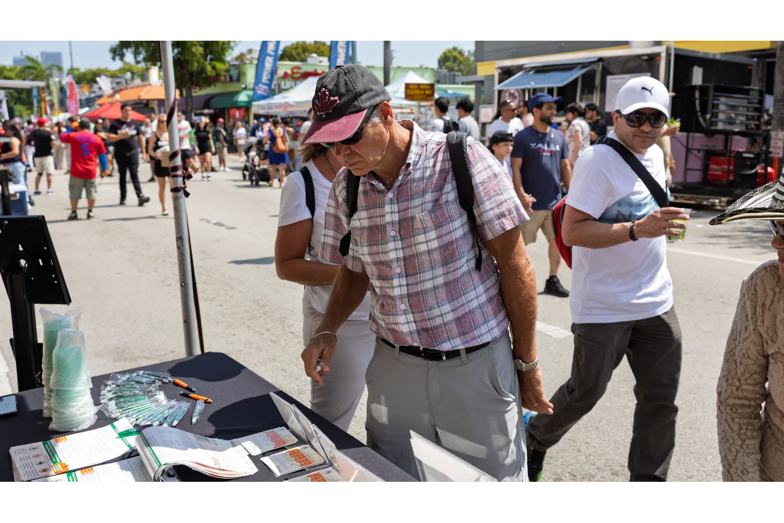 Man wearing a cap and plaid shirt browses a vendor booth at an outdoor street market on a sunny day.