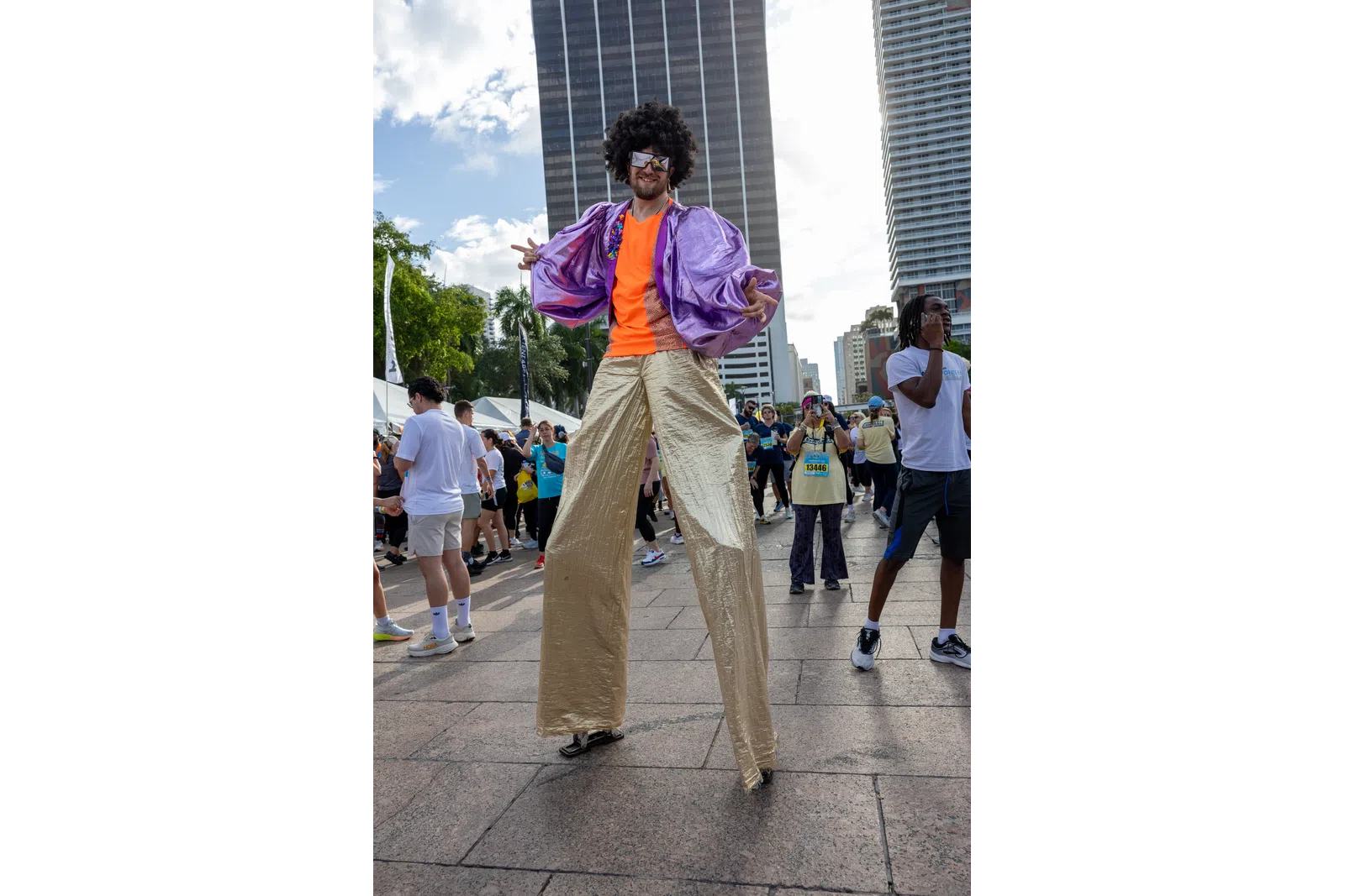Man on stilts wearing shiny purple jacket, orange shirt, gold pants, and afro wig at outdoor city event.