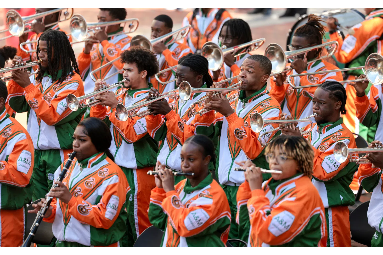 University marching band members in orange and green jackets playing brass and woodwind instruments during a performance.