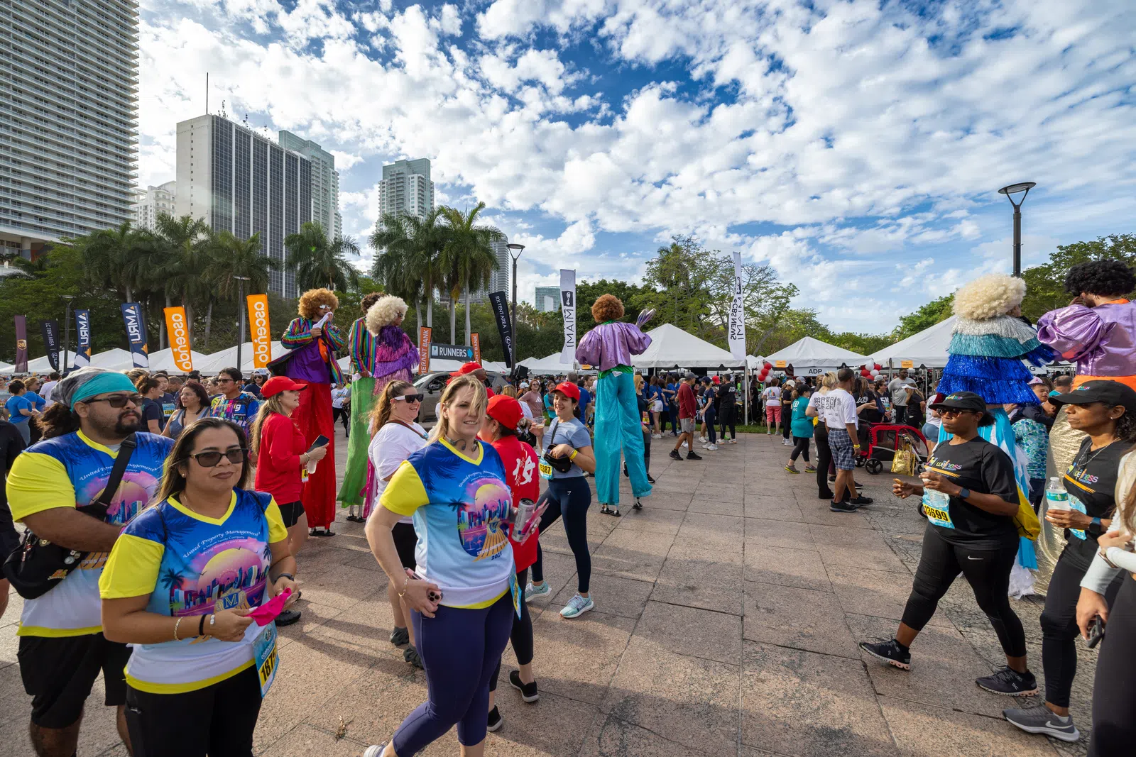 Crowd at outdoor event with performers on stilts, colorful costumes, tents, and modern buildings under a partly cloudy sky.