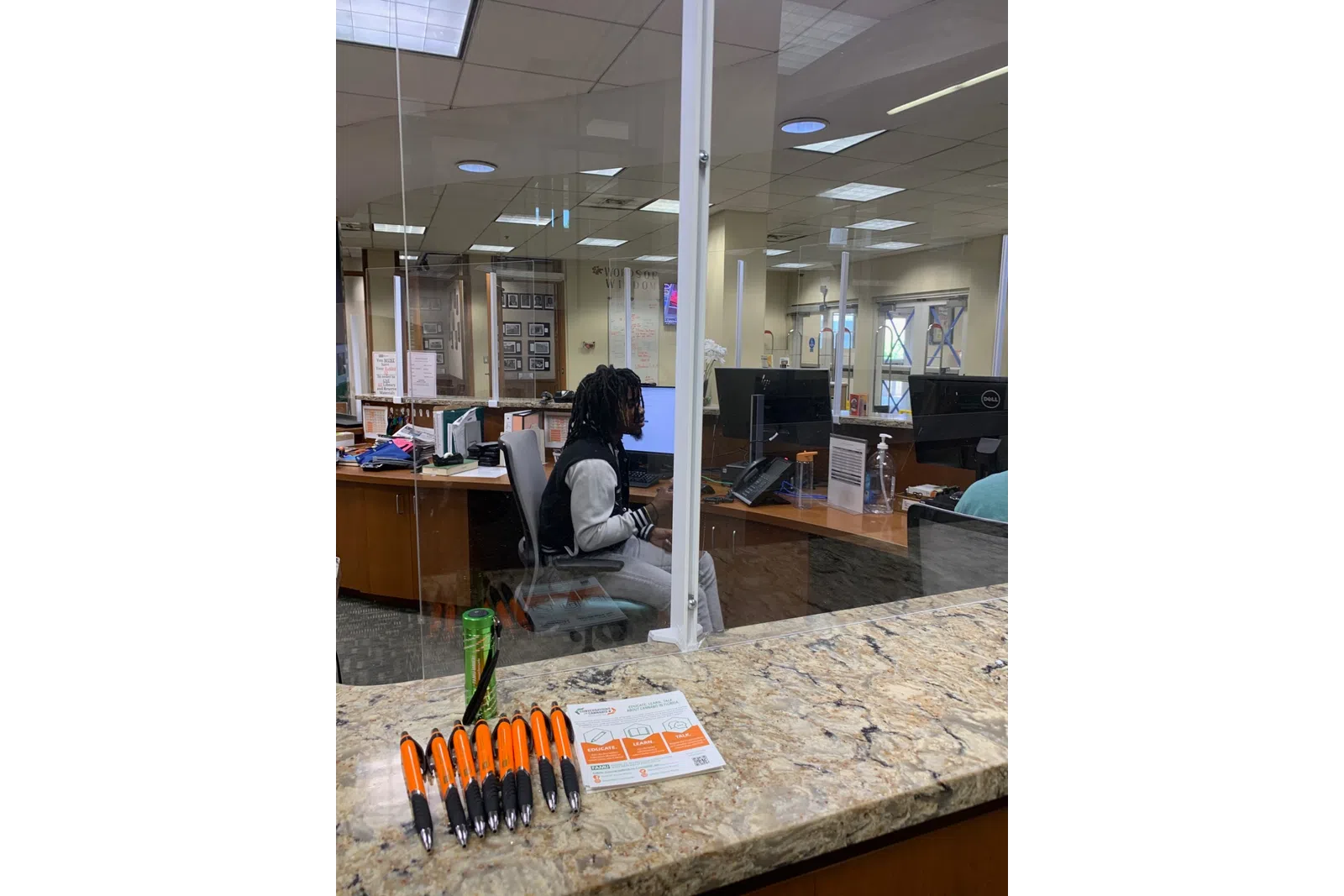 Office reception desk with pens, a water bottle, acrylic divider, and a person working on a computer.